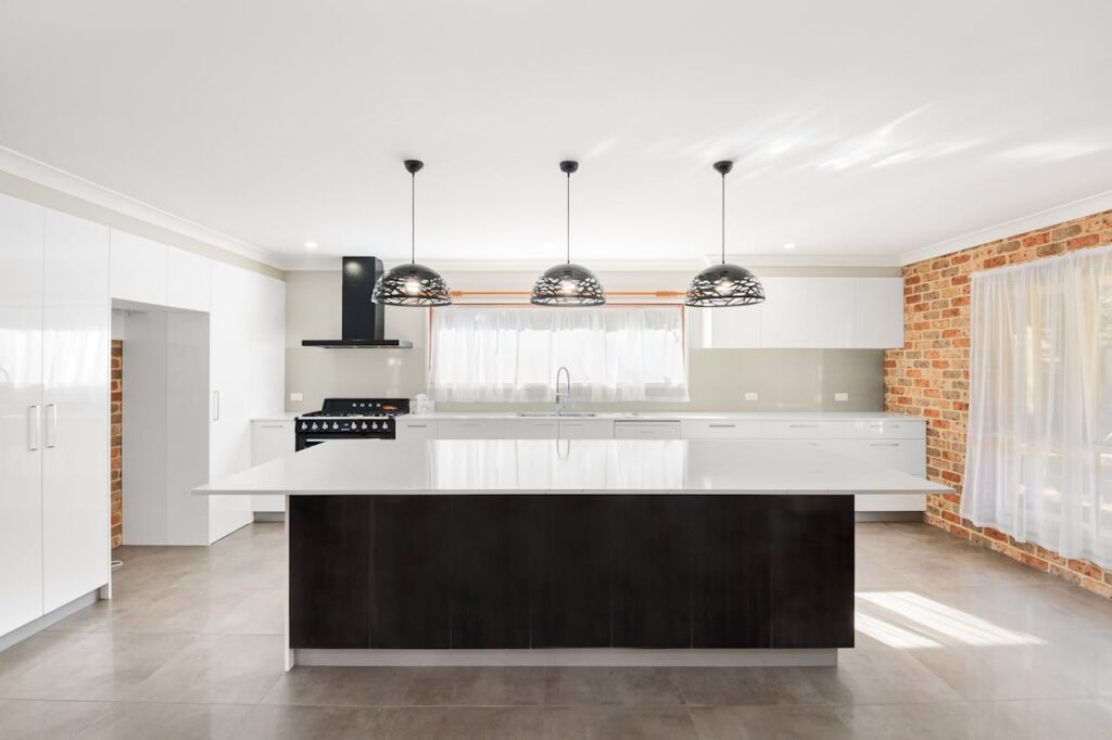 Spacious minimalist kitchen featuring white cabinets, pendant lighting, and brick walls.