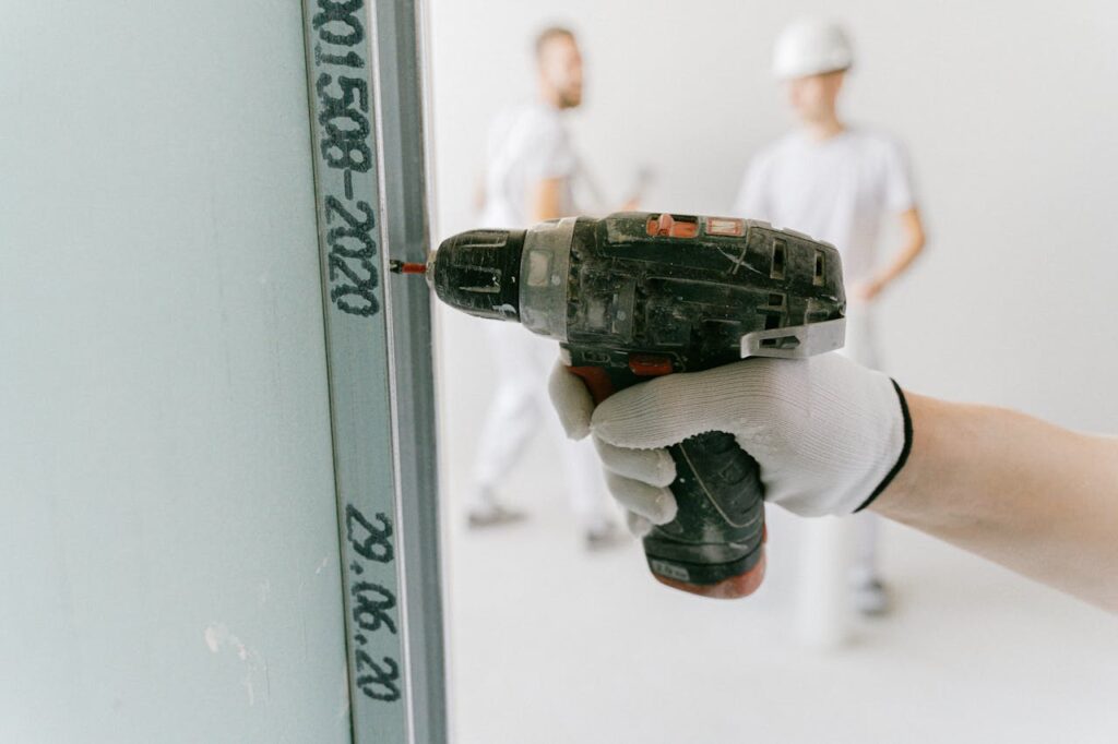 A construction worker uses a drill on drywall with blurred workers in the background.
